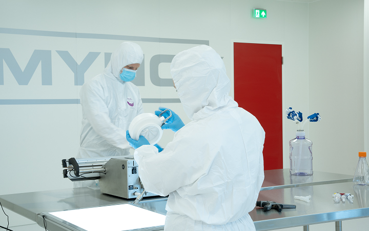 Two technicians wearing full cleanroom suits and gloves working in a laboratory setting.