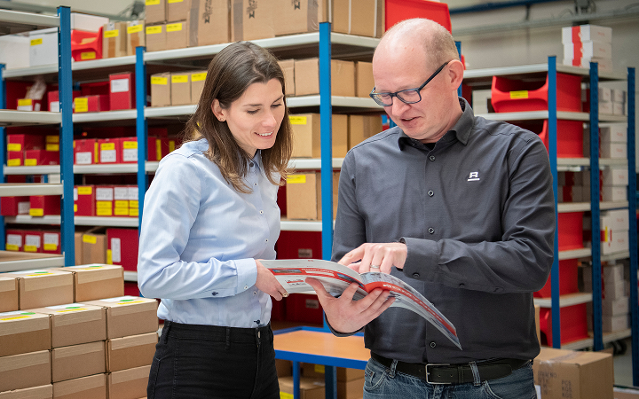 Two individuals in a warehouse discussing details from a catalog or document.