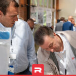 Two men inspecting a single-use system setup at an industrial exhibition, with a crowd in the background.