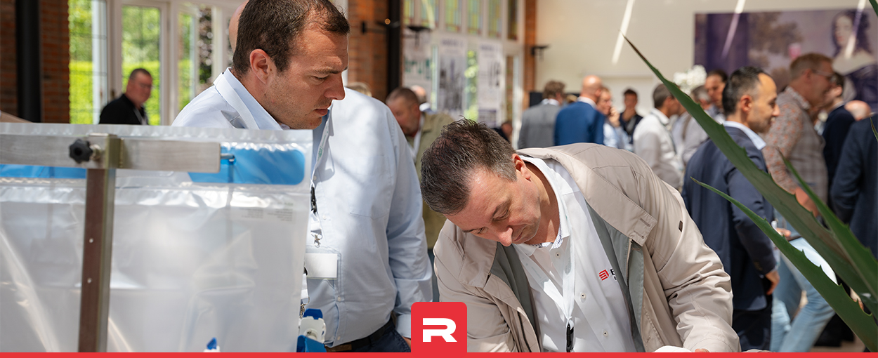 Two men inspecting a single-use system setup at an industrial exhibition, with a crowd in the background.